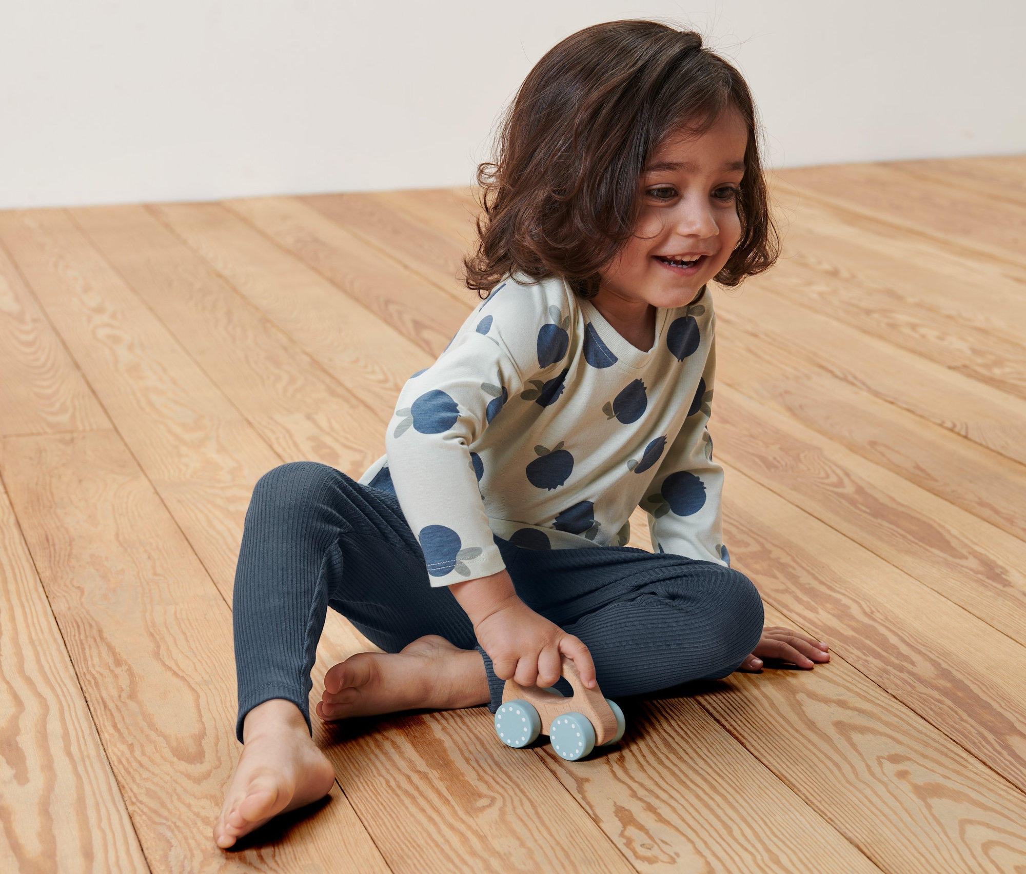 Ein Kind sitzt auf dem Boden und spielt mit einem kleinen Holzauto. Es trägt eine Kinder-Rippleggings.