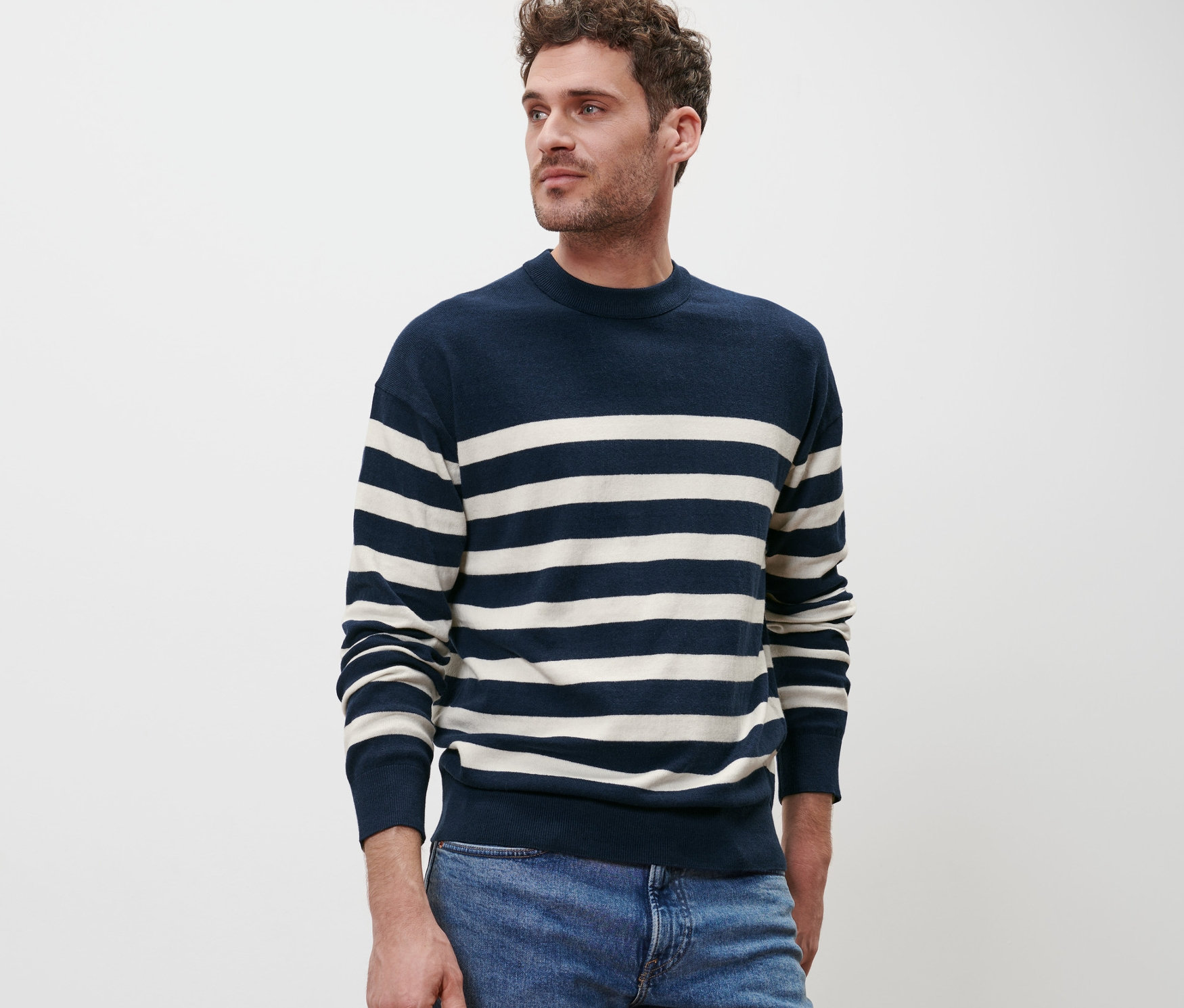 Mann posiert vor einem weißen Hintergrund in einem Pullover mit Rundhalsausschnitt mit blau-weißen Streifen und blauen Jeans.