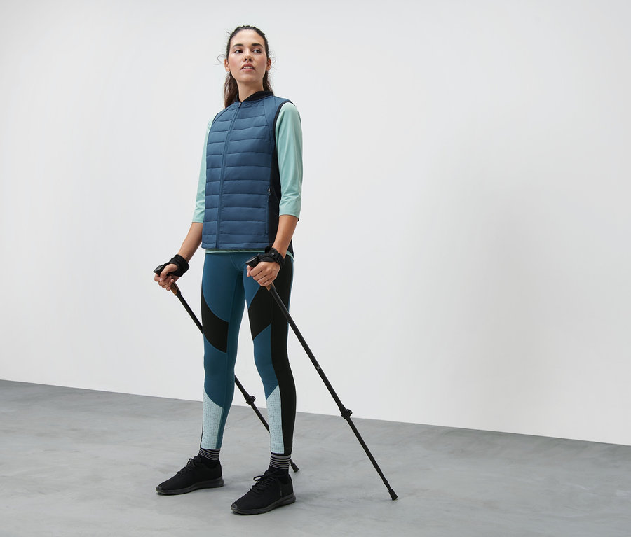 Frau hält Teleskop-Walkingstöcke. Sie trägt eine Thermo-Steppweste, eine Colorblocking-Sporttight und ein mintfarbenes Funktionsshirt mit 3/4-Ärmeln.