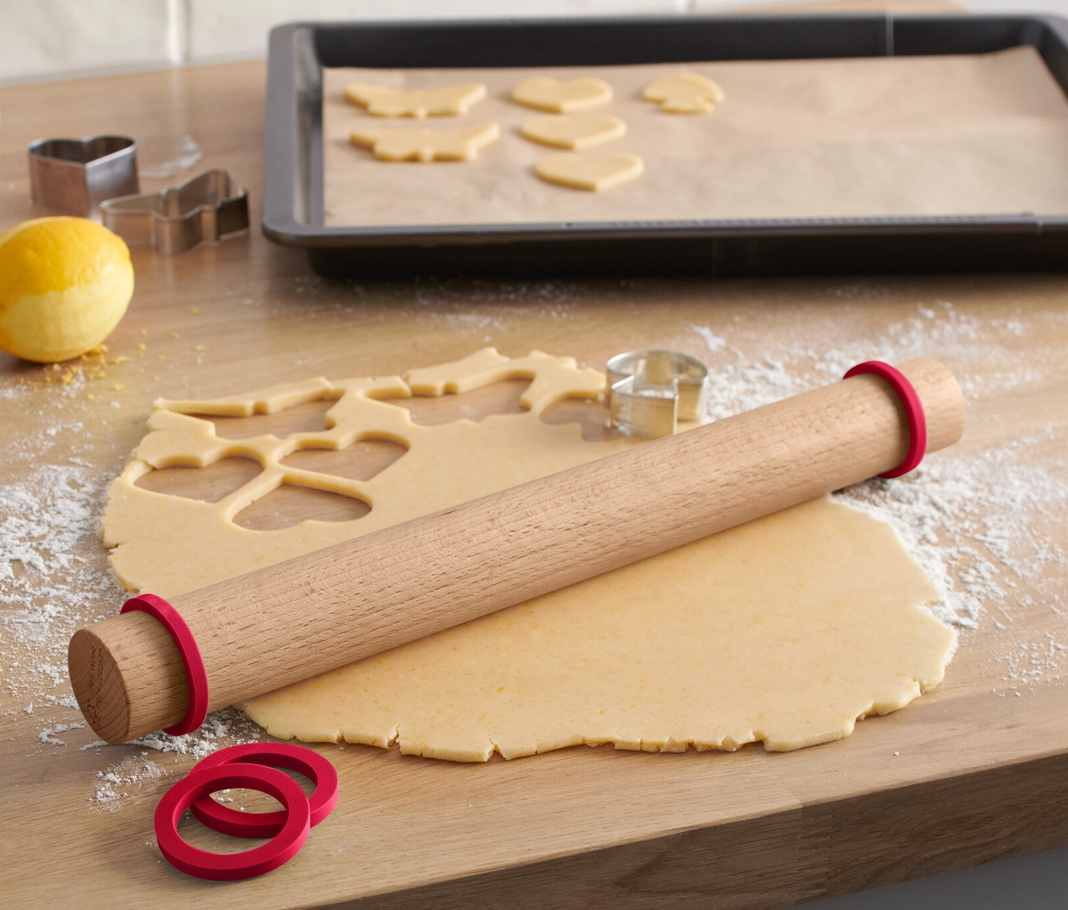 Auf einer Holzfläche liegen eine Holz-Teigrolle mit roten Gummiringen, daneben ausgerollter Teig mit Ausstechformen und Plätzchenformen.