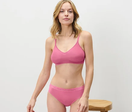 Eine blonde Frau in rosa Unterwäsche steht neben einem Holzhocker. Sie trägt 2 Slips, rosé.