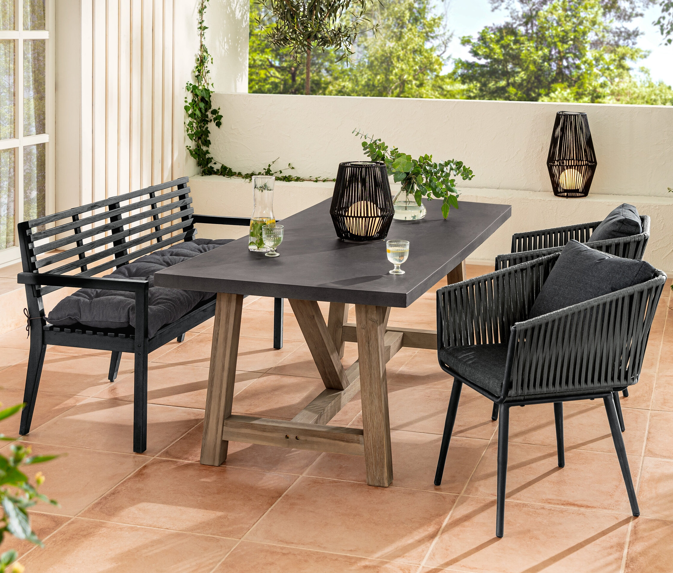 Eine Gartensitzgruppe mit einer Gartenbank mit Armlehne und einer Sun Garden Gartenbank-Auflage, einem Esstisch mit Polyzement-Tischplatte und Dining-Sesseln mit Textilgeflecht. Auf dem Tisch steht eine Solar-Laterne mit Textilband.