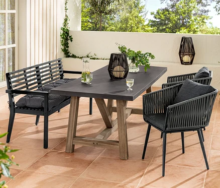 Eine Gartensitzgruppe mit einer Gartenbank mit Armlehne und einer Sun Garden Gartenbank-Auflage, einem Esstisch mit Polyzement-Tischplatte und Dining-Sesseln mit Textilgeflecht. Auf dem Tisch steht eine Solar-Laterne mit Textilband.