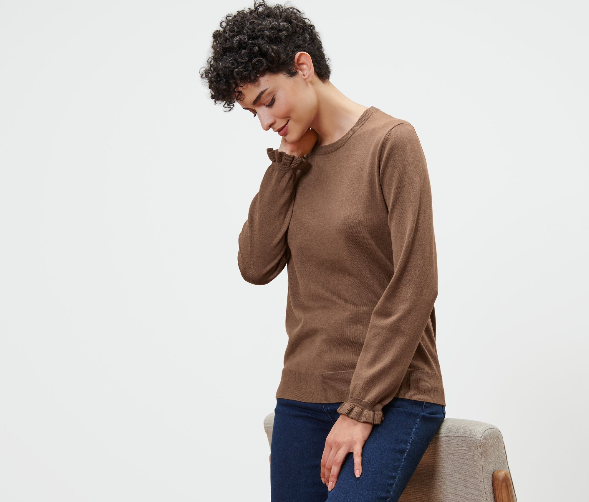 Frau lehnt an einem Stuhl, trägt einen braunen Feinstrickpullover mit Rüschendetail.