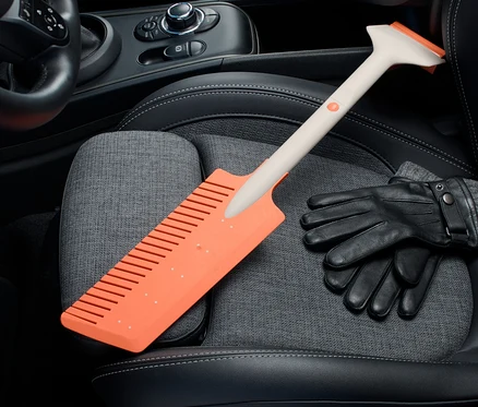 Ein orangefarbener Auto-Eiskratzer, steckbar, liegt auf einem Autositz neben schwarzen Handschuhen.