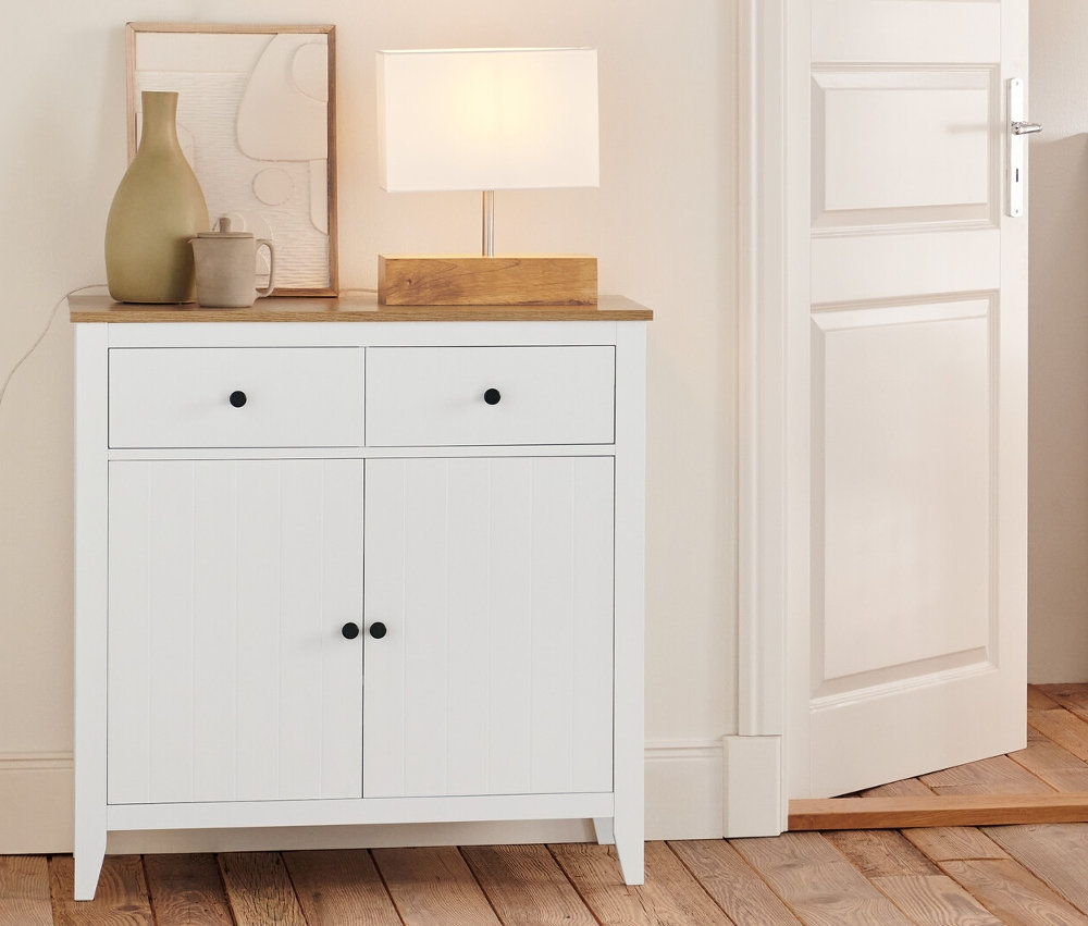 Im Raum stehen eine Kommode mit Schubladen, ein Sideboard, ein Regal und eine Tischleuchte mit Holzfuß.