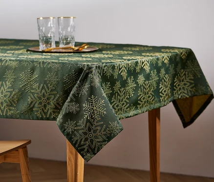 Auf einem Holztisch liegt eine grüne Jacquard-Tischdecke mit Schneeflockenmuster. Auf dem Tisch stehen Gläser und ein Holzstuhl.