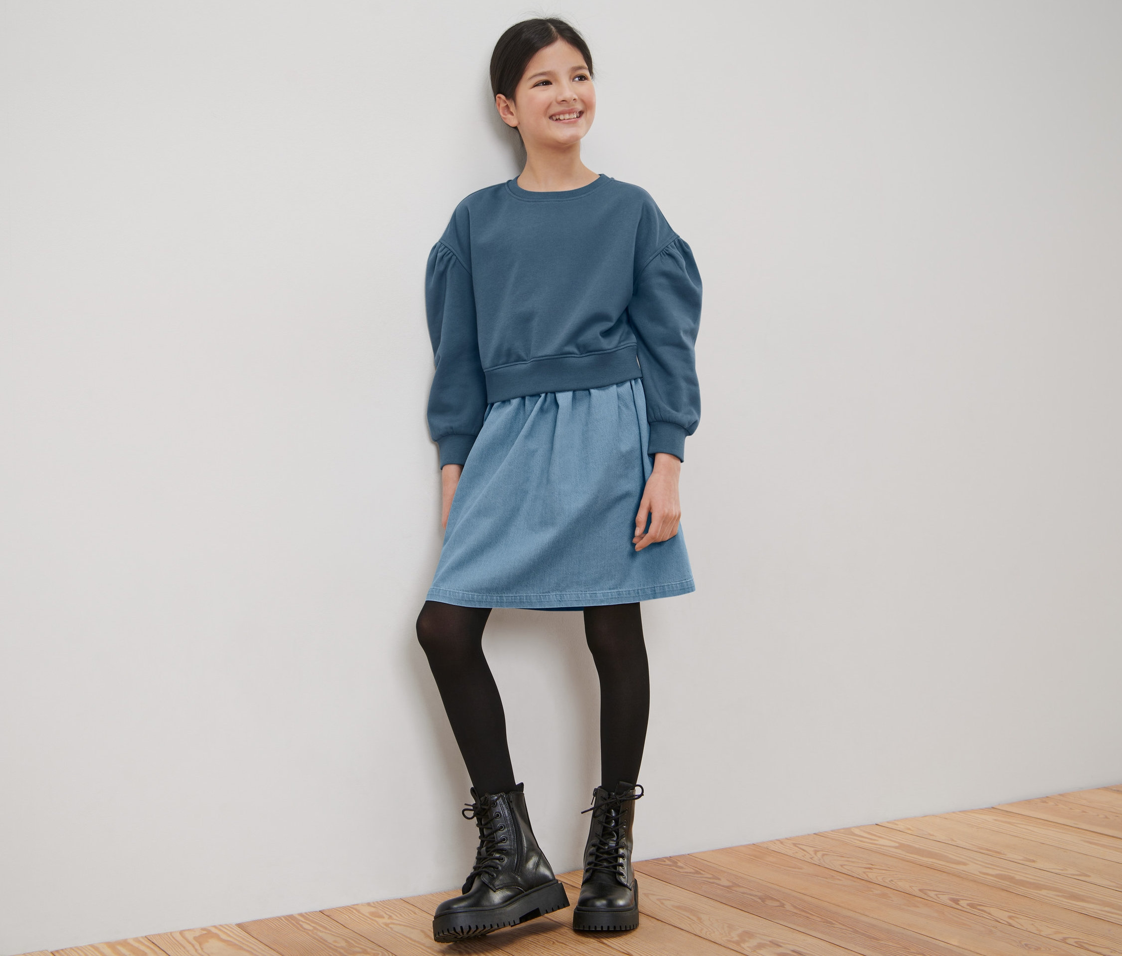 Ein lächelndes Mädchen im Kinder-Sweatkleid steht an einer weißen Wand.