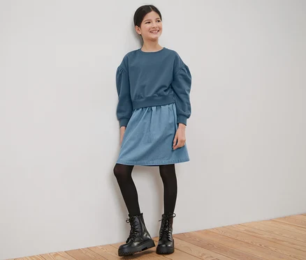 Ein lächelndes Mädchen im Kinder-Sweatkleid steht an einer weißen Wand.