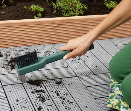 Frau reinigt mit einer Garten-Fugen-Reinigungsbürste die Fugen zwischen den Fliesen. Im Hintergrund befindet sich ein Blumenbeet.