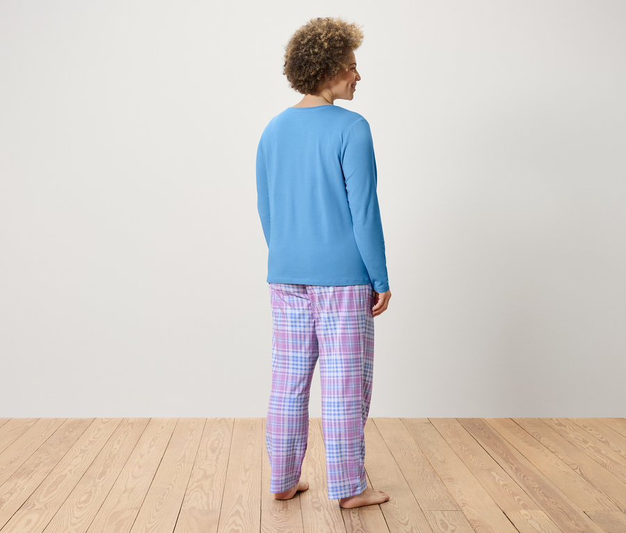 Frau mit lockigem Haar steht mit dem Rücken zur Kamera und trägt ein blaues Langarmshirt und eine karierte Hose.
