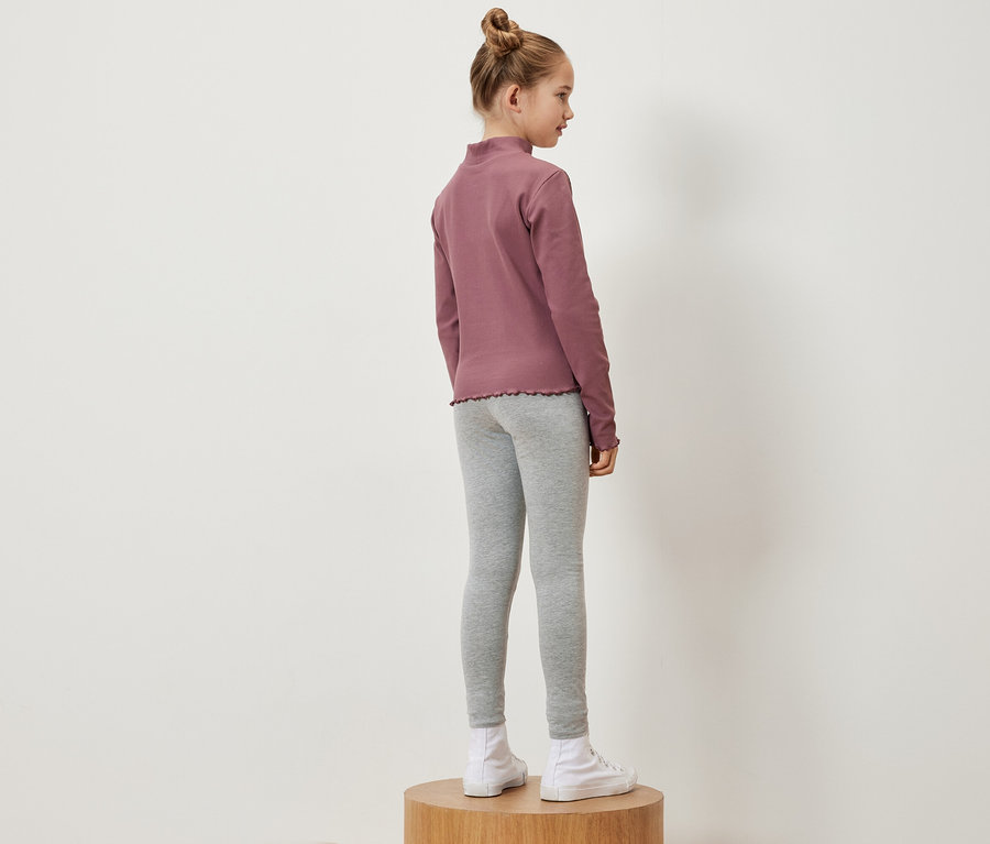 Mädchen steht seitlich auf einem Holzsockel und trägt ein rosafarbenes Langarmshirt, graue Leggings und weiße Turnschuhe.