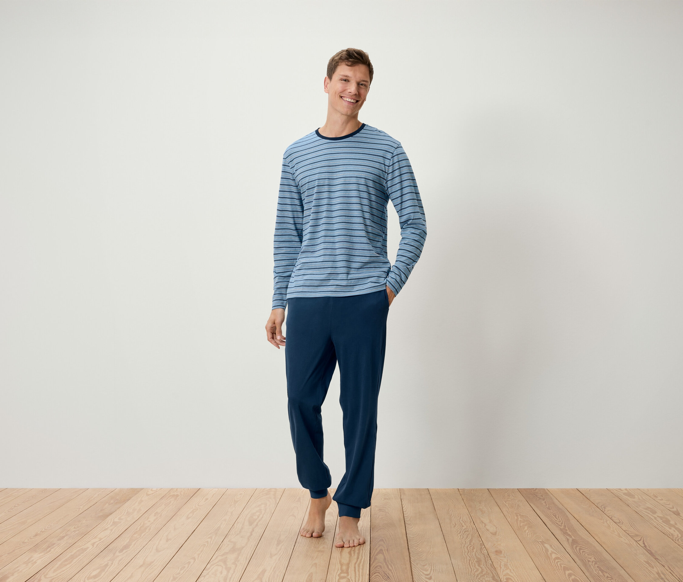 Mann steht barfuß auf Holzboden, trägt einen hellblau gestreiften Pyjama mit Bündchen.