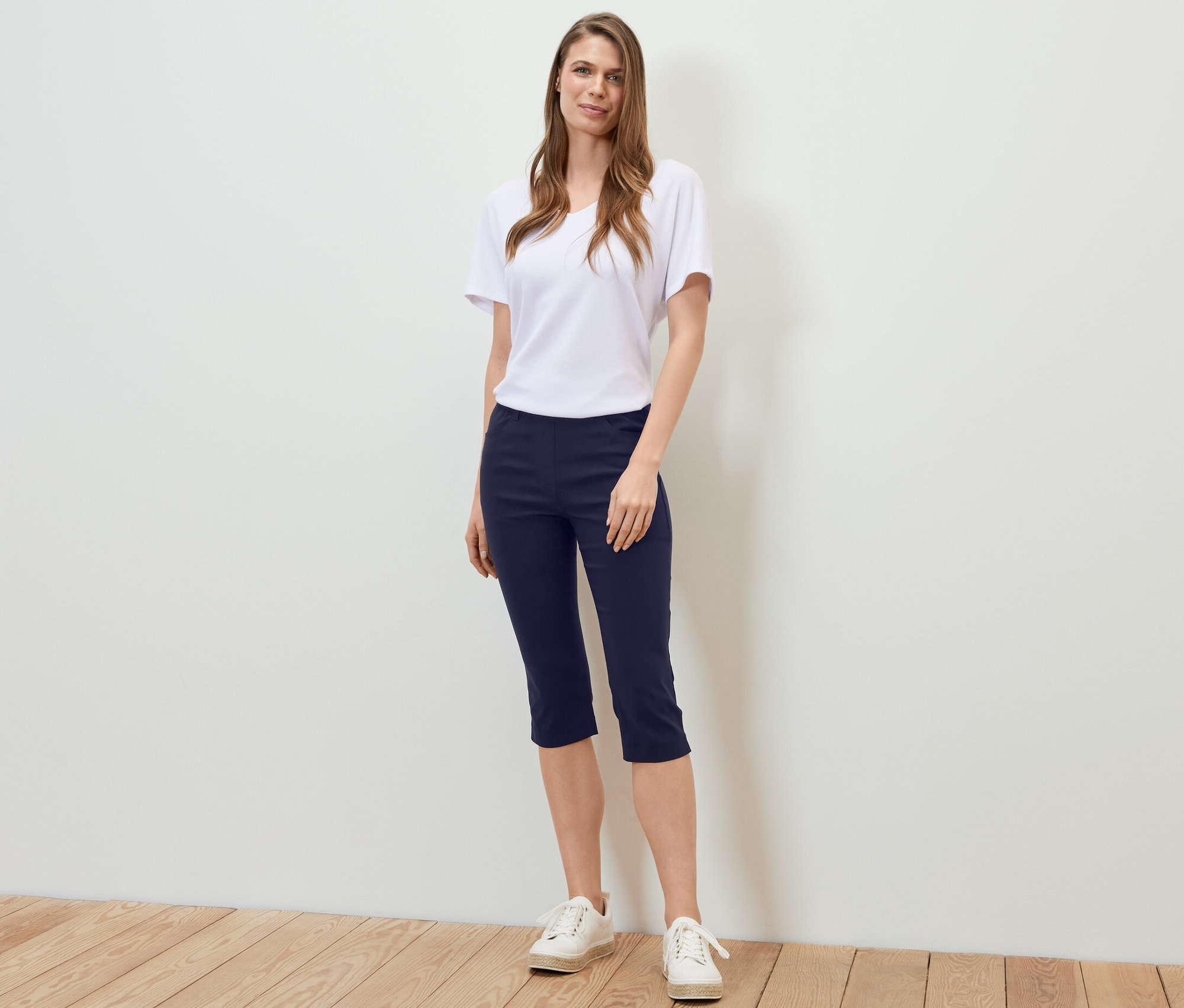 Frau steht und trägt eine dunkelblaue 3/4-Bengalin-Hose, ein weißes T-Shirt und Turnschuhe.