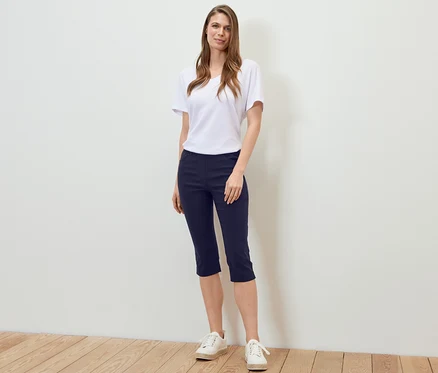 Frau steht und trägt eine dunkelblaue 3/4-Bengalin-Hose, ein weißes T-Shirt und Turnschuhe.