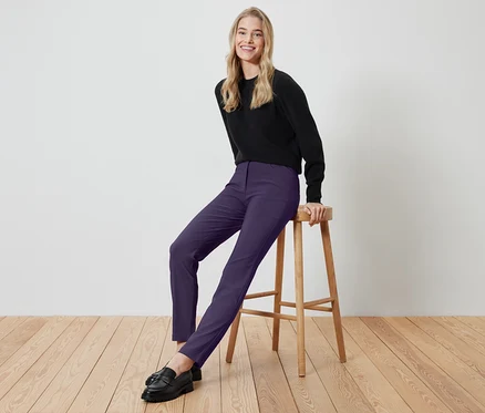 Eine blonde Frau sitzt auf einem Holzhocker und trägt ein schwarzes Oberteil und violette Hosen.