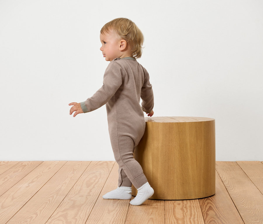 Kleinkind in braunem Overall lehnt an einem Holzhocker.