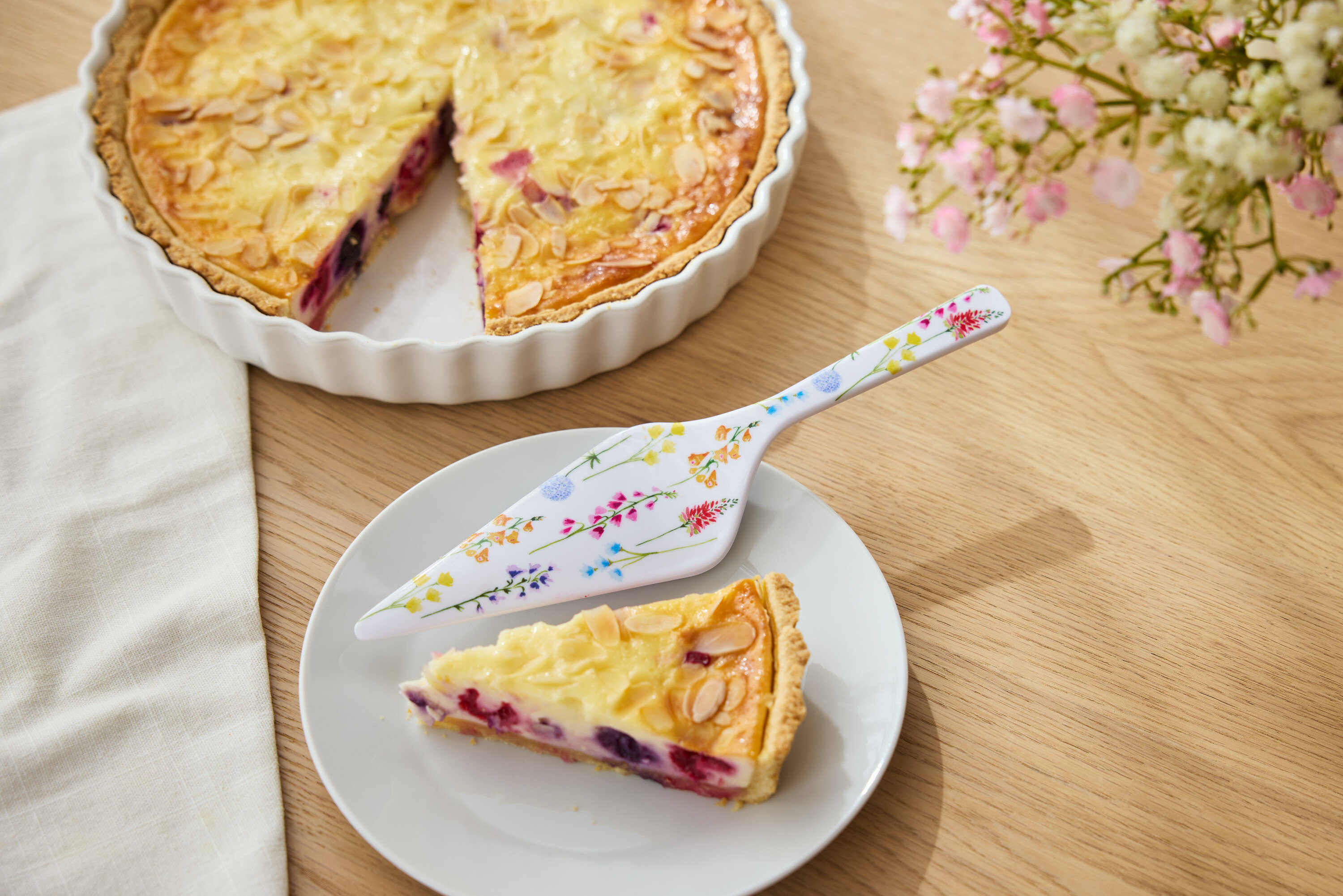 Obstkuchen mit Mandeln in einer weißen Form, ein Stück Kuchen auf einem Teller und ein Tortenheber mit Blumenmuster.