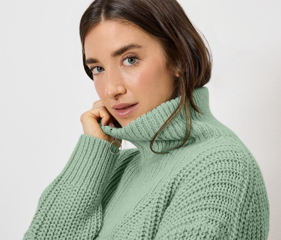Frau mit hellbraunem Haar und hellgrünem Rollkragenpullover posiert und schaut in die Kamera.