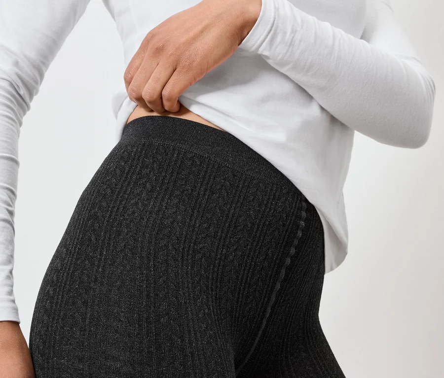Frau in grauen Strickleggings und weißem Langarmshirt, die den Bund der Leggings anpasst.
