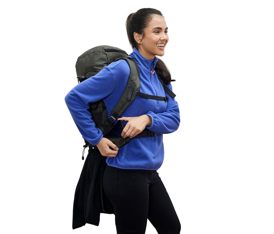 Eine lächelnde Frau in einer blauen Fleecejacke justiert den Gurt an einem grünen 40-l-Wander-Rucksack.