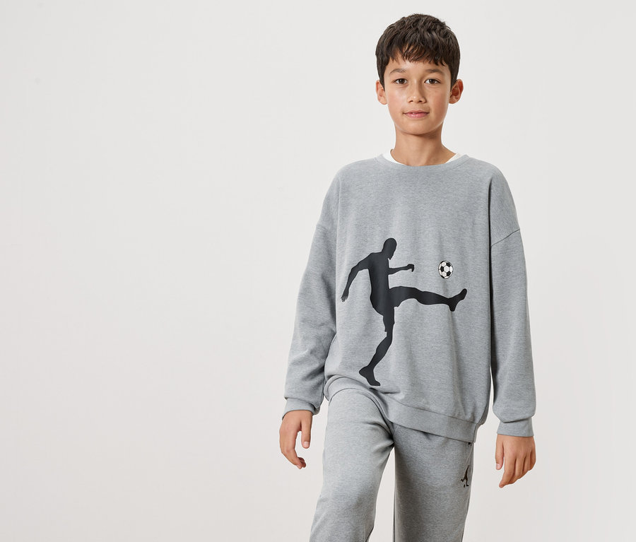 Ein Junge trägt einen grauen Pullover mit einer schwarzen Silhouette eines Fußballspielers und einem Ball darauf, sowie graue Jogginghosen.