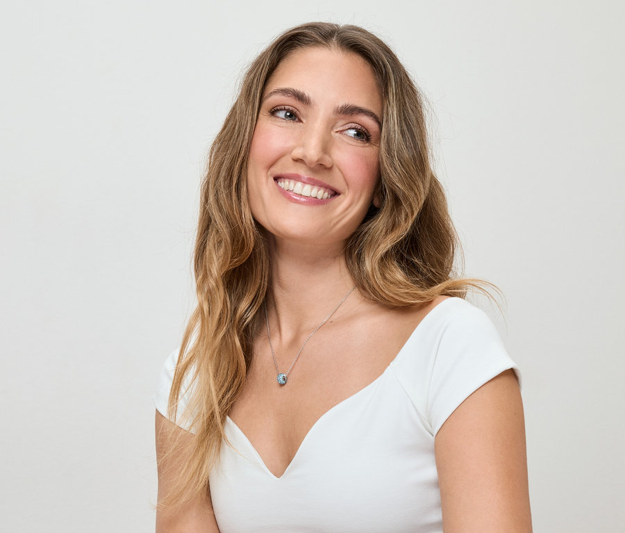Frau mit gewelltem, braunem Haar und weißem Oberteil lächelt und trägt eine silberne Halskette mit blauem Anhänger.