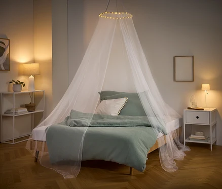 Ein Bett mit grüner Bettwäsche und einem weißen Moskitonetz mit Lichtern hängt von der Decke. Nachttisch und Regal stehen an den Seiten.
