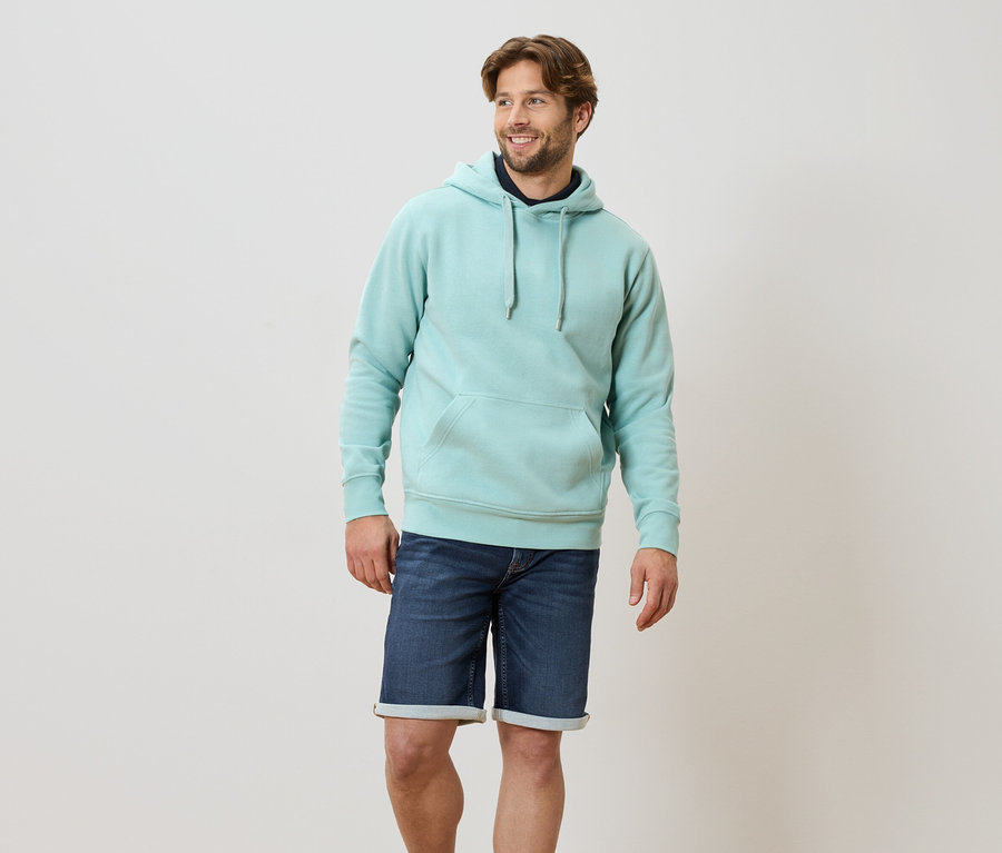 Ein Mann steht und trägt einen helltürkisfarbenen Hoodie und blaue Jeansshorts.