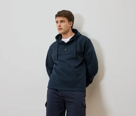 Mann mit dunkelblauem Kapuzenpullover und Shorts vor einer weißen Wand.