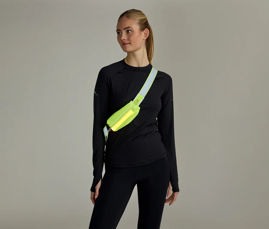 Frau mit Pferdeschwanz, schwarzem Langarmshirt und Leggings, trägt eine neongelbe Bauchtasche quer über der Brust.