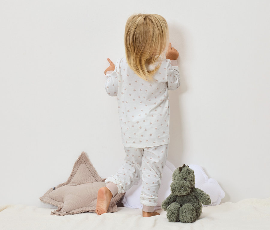 Ein Kind in einem Sternenpyjama steht mit dem Gesicht zur Wand, daneben ein Sternkissen und ein grünes Stofftier.