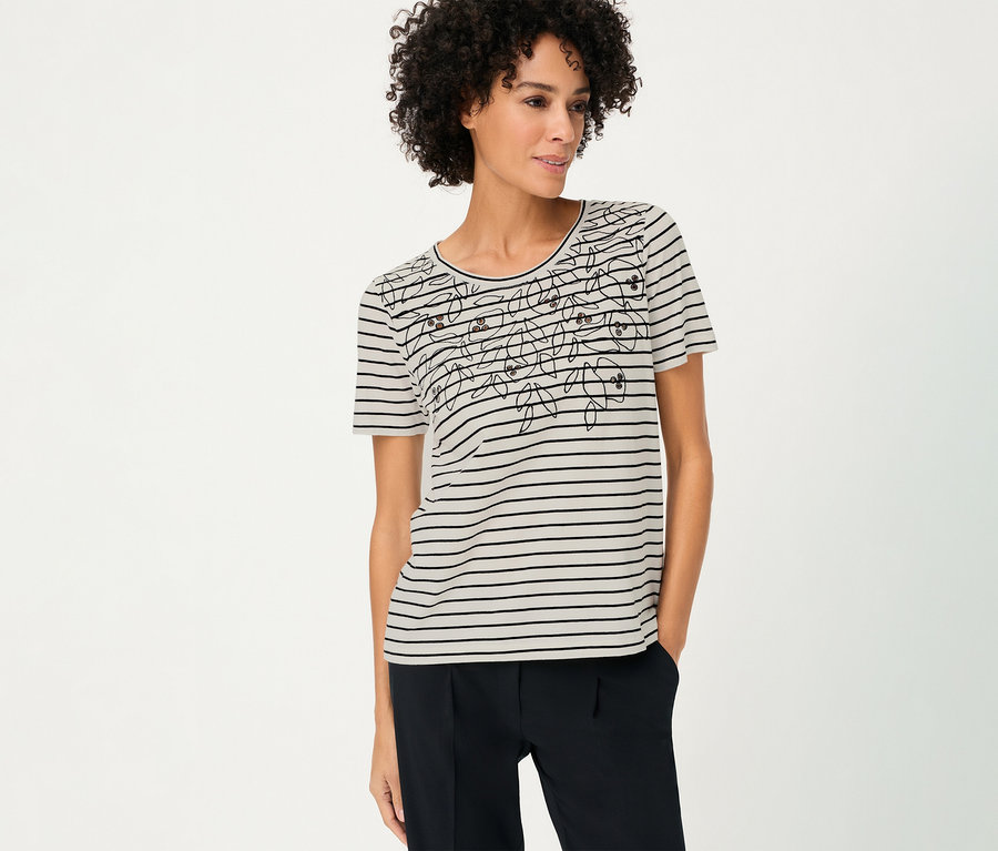 Frau mit lockigem Haar trägt ein gestreiftes Olsen Shirt mit Blumenmuster und dunkle Hose.