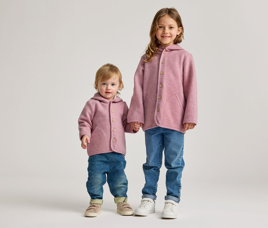 Zwei Kinder, ein Kleinkind und ein älteres Mädchen, stehen nebeneinander und halten Händchen. Beide tragen rosa Kapuzenjacken mit Holzknöpfen und blaue Jeans. Das Kleinkind trägt beige Schuhe, das Mädchen weiße Sneaker.