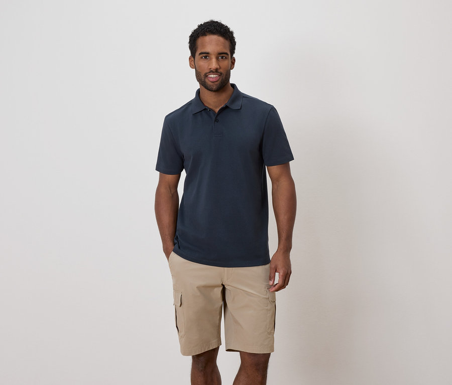 Ein Mann mit dunklen Haaren und Bart trägt ein marineblaues Poloshirt und Khaki-Cargo-Shorts.