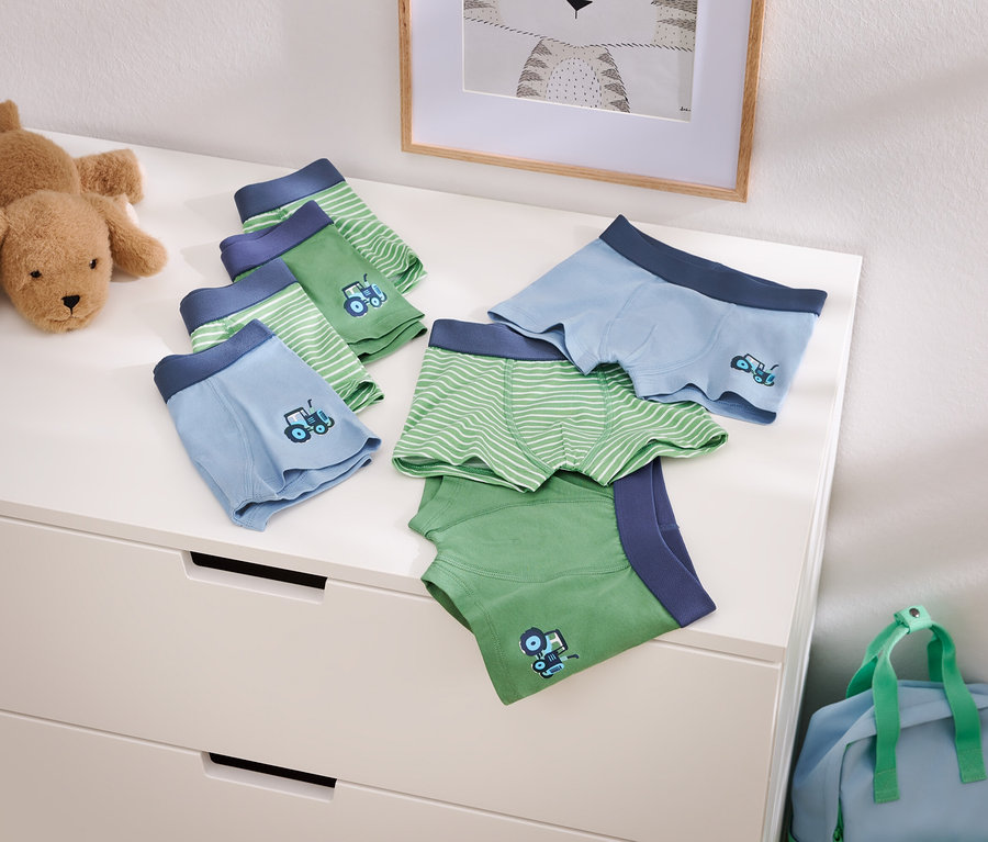 Abbildung von hellblauen und grünen Jungenunterhosen mit Traktormotiv auf einer Kommode, neben einem Stofftierhund und einer blauen Reisetasche.