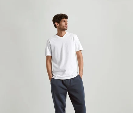 Mann mit lockigem Haar, der ein weißes V-Ausschnitt-T-Shirt und marineblaue Jogginghose trägt.