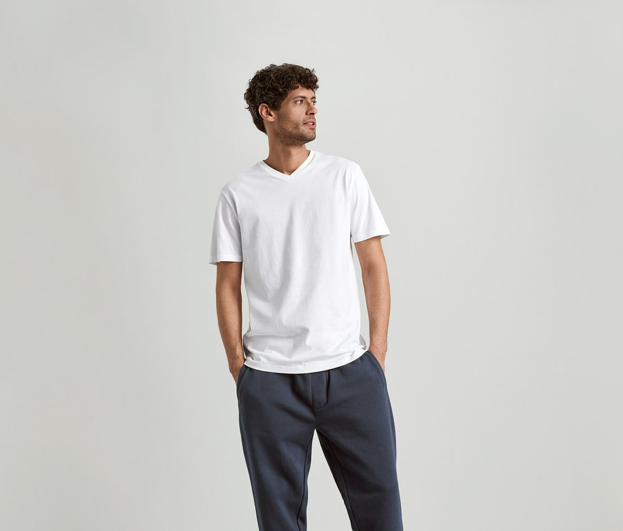 Mann mit lockigem Haar, der ein weißes V-Ausschnitt-T-Shirt und marineblaue Jogginghose trägt.