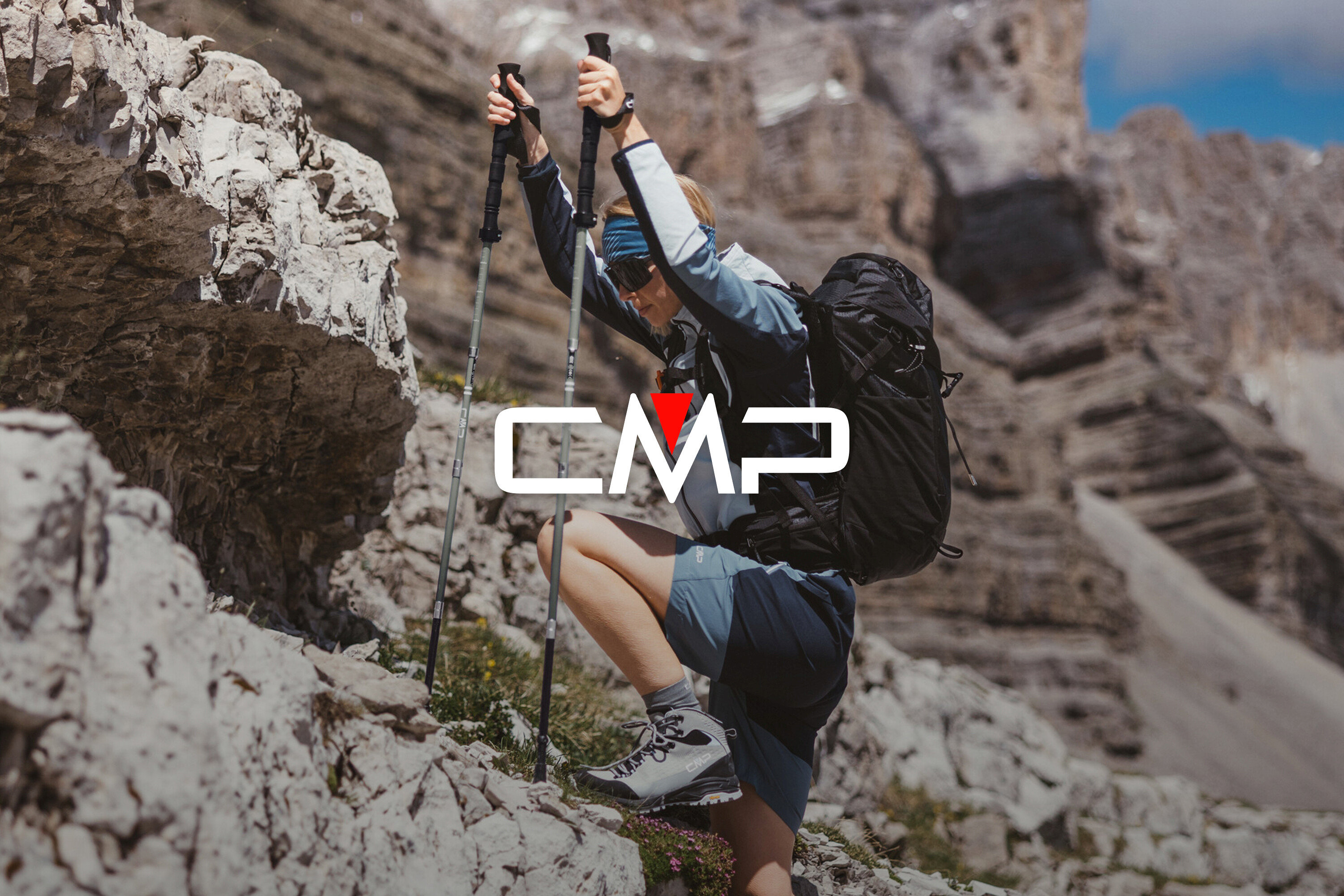 Frau mit Rucksack und Wanderstöcken klettert auf Felsen. Sie trägt Wanderschuhe, Shorts und eine Jacke.