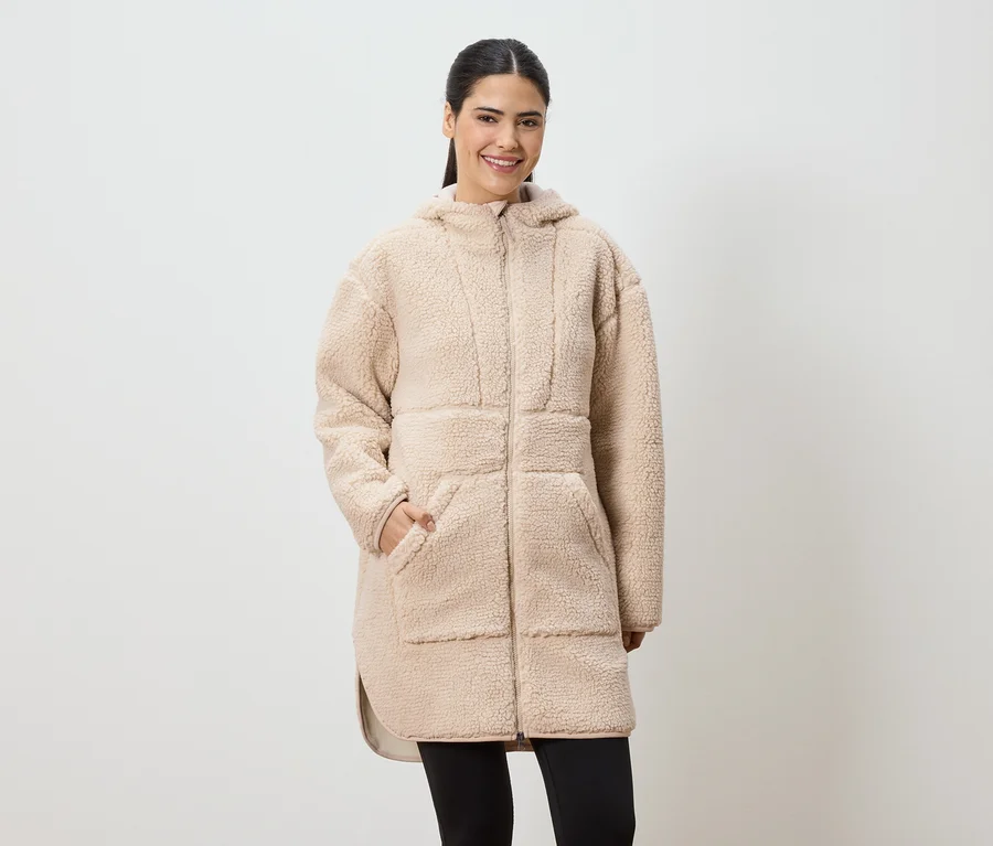 Frau mit Pferdeschwanz trägt einen langen, beigen Teddyfellmantel mit Kapuze und schwarzen Leggings.