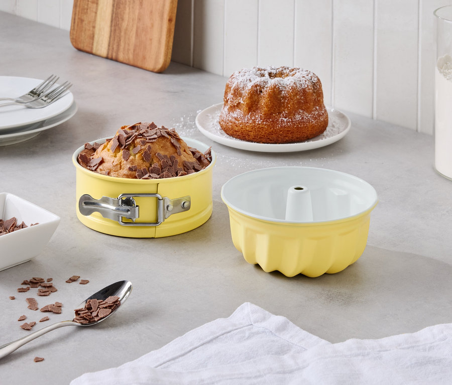 Ein gedeckter Tisch mit einem Gugelhupf Kuchen mit Puderzucker, einem Kuchen mit Schokoladenstückchen in einer gelben Springform, einer leeren gelben Gugelhupfform und einem Glas mit Mehl.