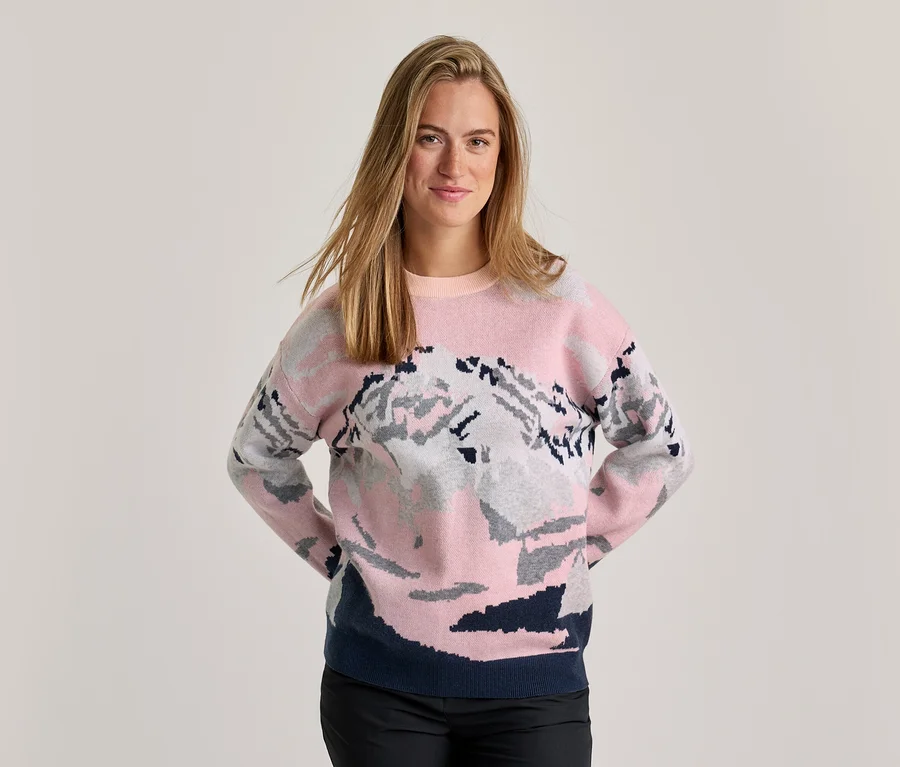 Lächelnde Frau in einem rosa-grau-dunkelblauen Pullover mit Bergmuster, schwarzer Hose und Händen hinter dem Rücken.