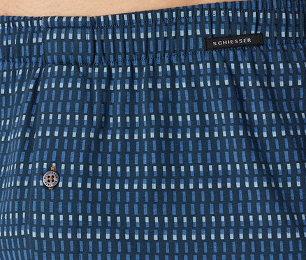 Nahaufnahme eines blauen Stoffes mit Muster aus hell- und dunkelblauen Rechtecken, oben ein elastischer Bund, schwarzes Etikett mit „SCHIESSER“-Schriftzug und ein Knopf mit „SCHIESSER EST. 1875“.