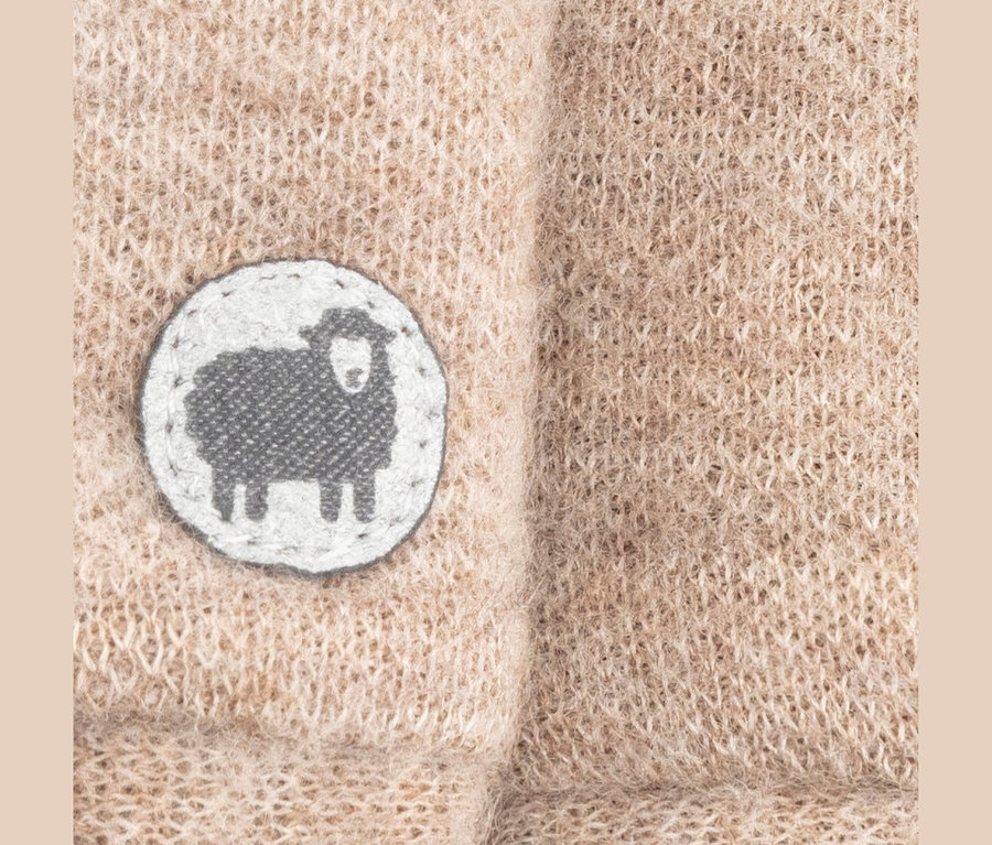 Nahaufnahme eines hellbraunen Strickstoffs mit einem runden weißen Aufnäher, der eine graue Schafsilhouette zeigt.