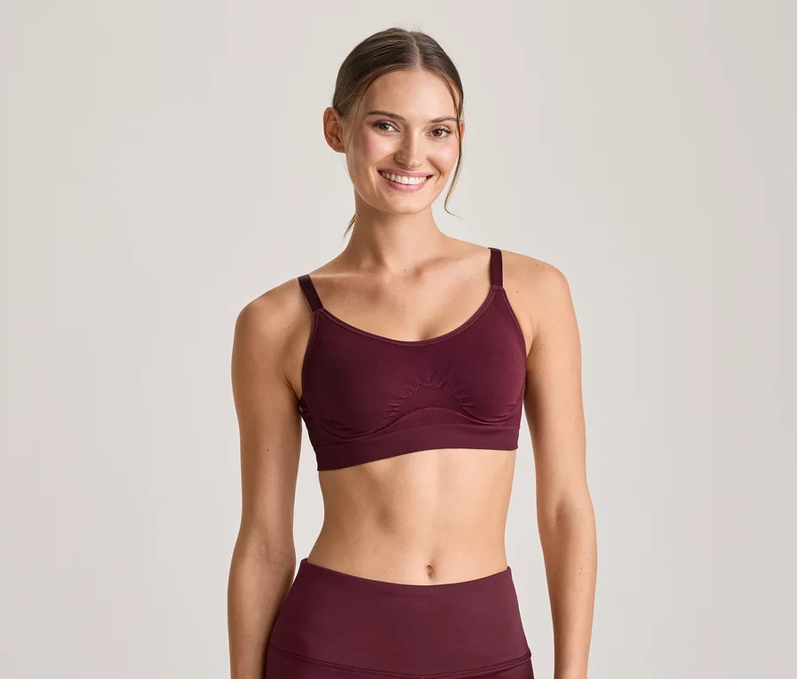 Lächelnde Frau in einem bordeauxroten Sport-BH und passenden Leggings, blickt in die Kamera.