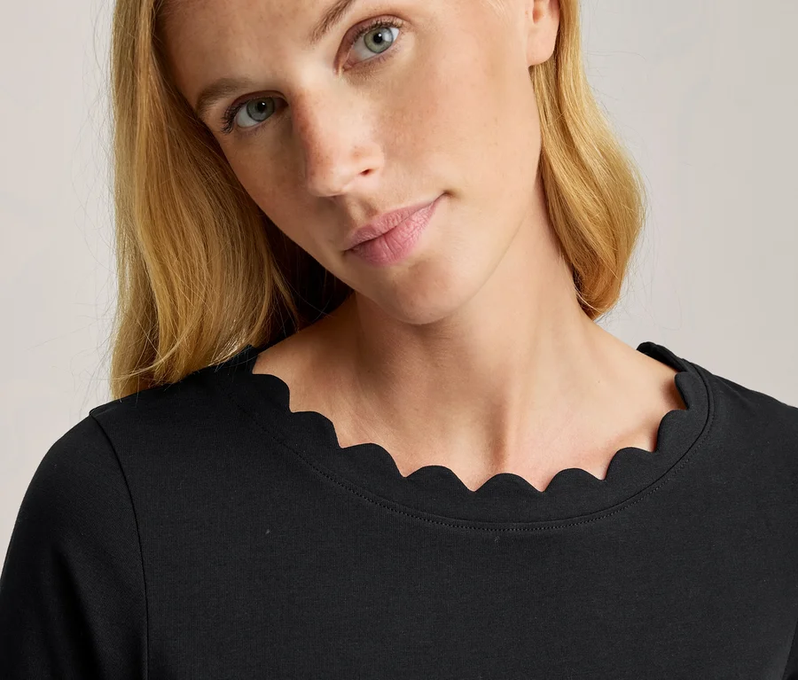 Nahaufnahme einer Frau mit blondem Haar und blauen Augen, die ein schwarzes Langarmshirt mit Wellenkante trägt.