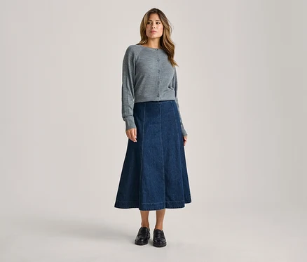 Eine Frau steht da und trägt einen grauen Strickcardigan, einen dunkelblauen Jeans-Midi-Rock in A-Linie und schwarze Loafer.