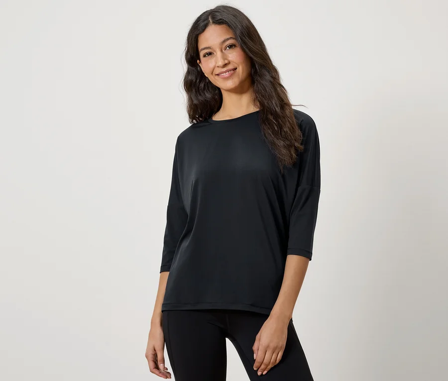 Eine Frau mit braunen Haaren posiert in einem schwarzen Dreiviertelarm-T-Shirt und schwarzen Leggings.