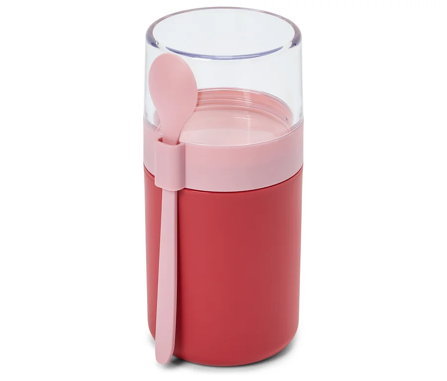 Ein roter Lebensmittelbehälter mit einem rosafarbenen Band und einem transparenten Deckel, an dessen Seite ein rosafarbener Löffel befestigt ist.