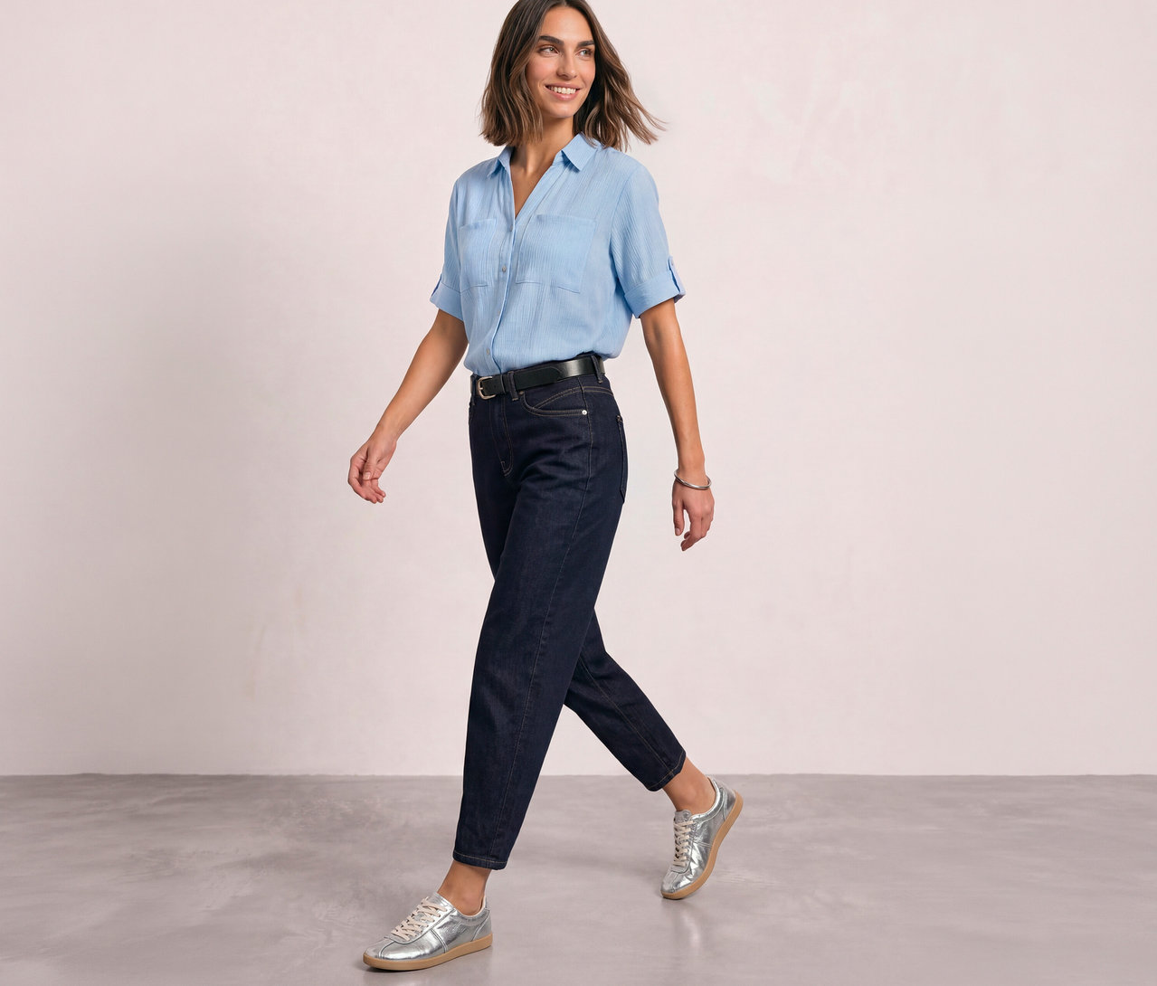Eine Frau geht in einer hellblauen Bluse, einem schwarzen Gürtel und einer Street One Barrel Jeans in Dunkelblau mit silbernen Sneakern.